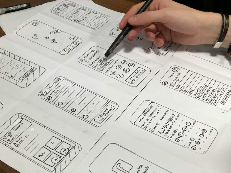 Main pointant un prototype d’application mobile dessiné à la main, avec différentes interfaces utilisateur schématisées. Image illustrant un article sur comment créer une interface utilisateur intuitive, centrée sur l’expérience et l’ergonomie